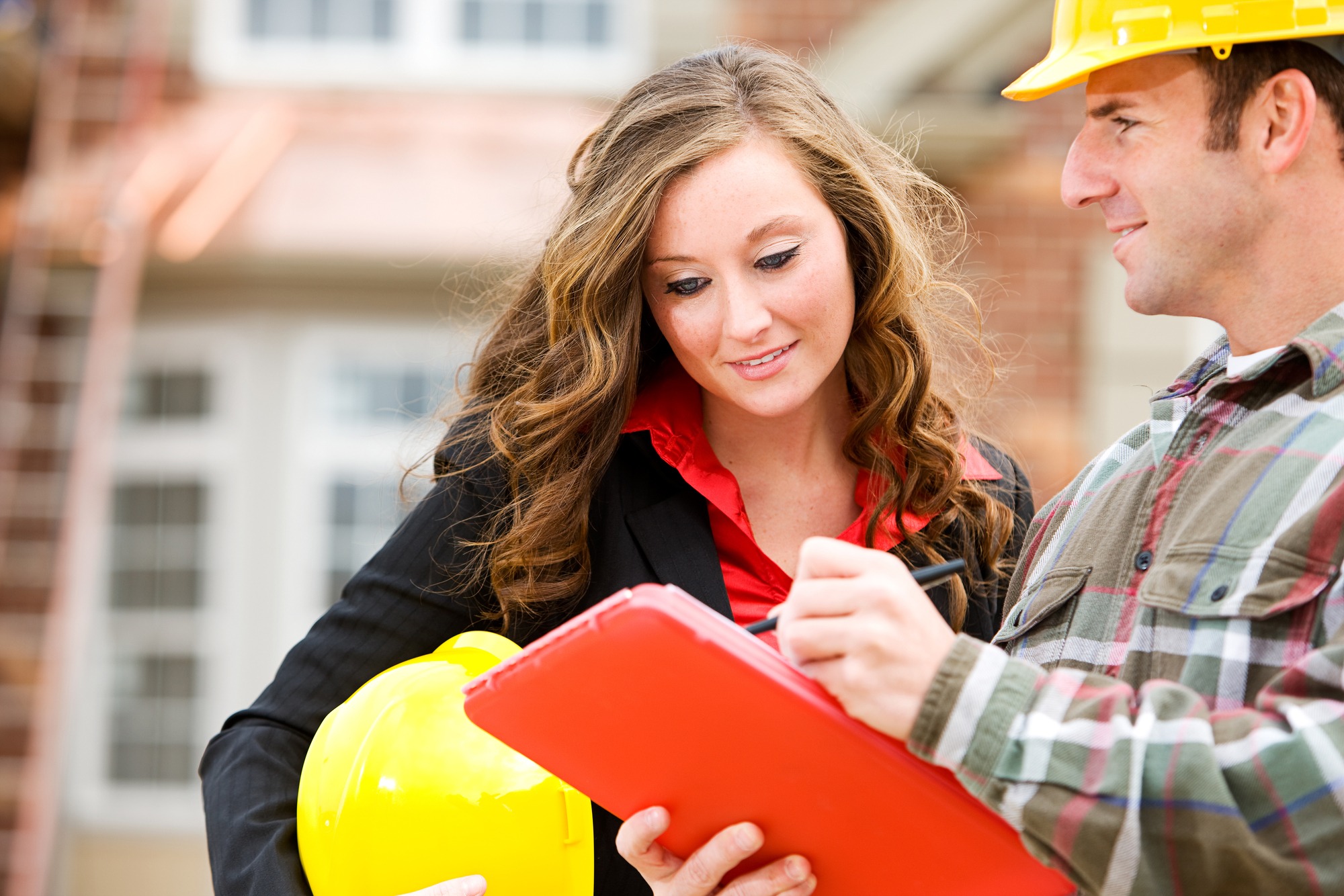 A female professional in a black blazer holds a yellow hard hat while discussing plans with a male construction worker in a plaid shirt.